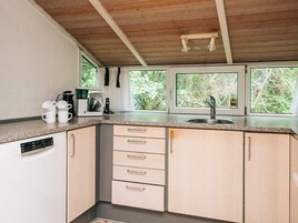 A compact kitchen with a built-in dishwasher, a coffee maker, and a sink. There are three windows providing natural light and a view of greenery outside.