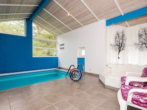 Une piscine intérieure à l’eau d’un bleu cristallin, équipée d’une bouée de sauvetage et dotée d’un espace détente blanc avec vue sur l’extérieur par une fenêtre.