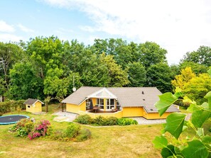 Une maison jaune avec un porche abrité, un petit abri de jardin et un trampoline, le tout dans un jardin verdoyant et luxuriant.