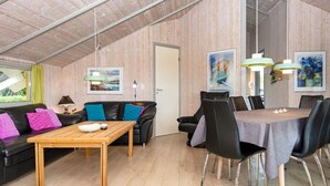 A cozy room with a wooden ceiling, a black leather sofa with colorful cushions, a wooden table, and a dining area with chairs and a table.