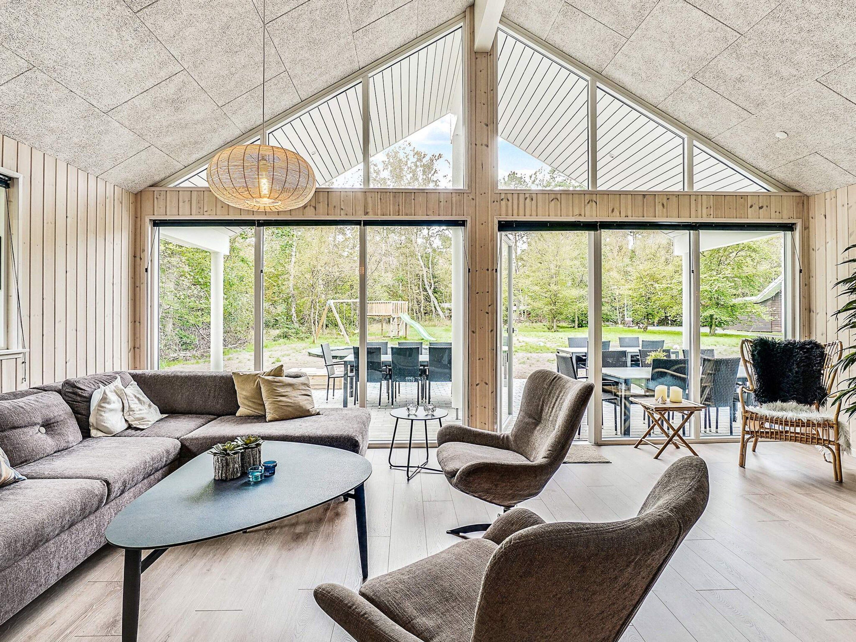 A spacious room with a high ceiling, large windows, a wooden interior, a sofa, a coffee table, and a dining area with chairs.