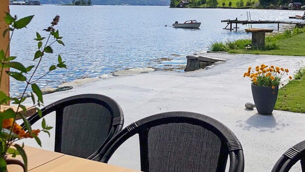 Eine Terrassensitzgelegenheit am Seeufer mit Korbstühlen und einem Tisch, mit Blick auf einen ruhigen See mit Booten und Häusern im Hintergrund.
