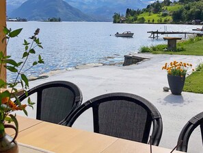 A lakeside patio with wicker chairs and a table, overlooking a calm lake with boats and houses in the background. - 6 Person Holiday Home in Oystese (Kvam)