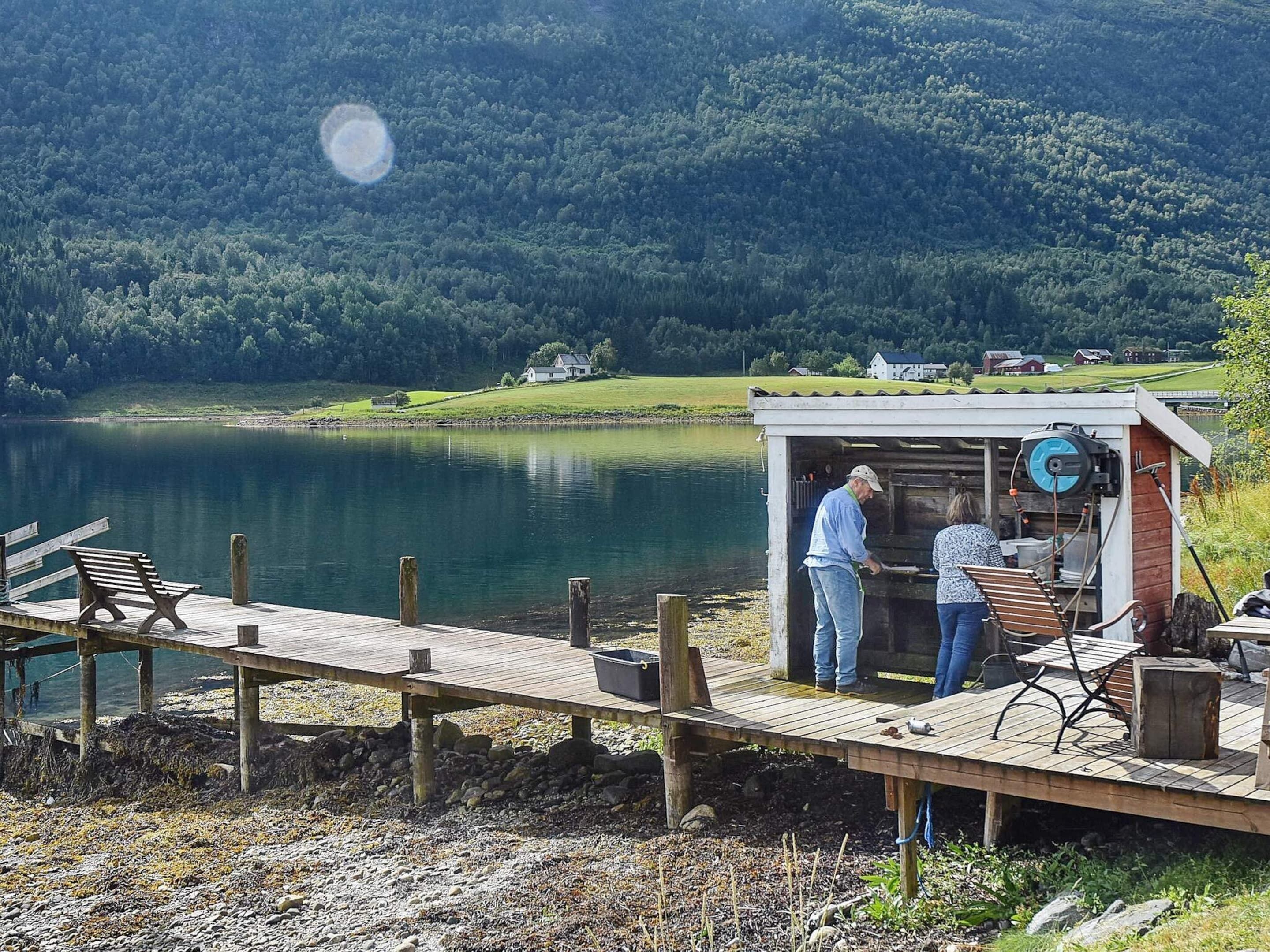 A wooden dock with a bench, a small wooden structure with a water pump, and a lake with houses in the background.