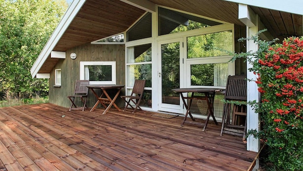 Um deck de madeira com uma mesa, cadeiras, uma pequena janela e uma porta que dá para um jardim.