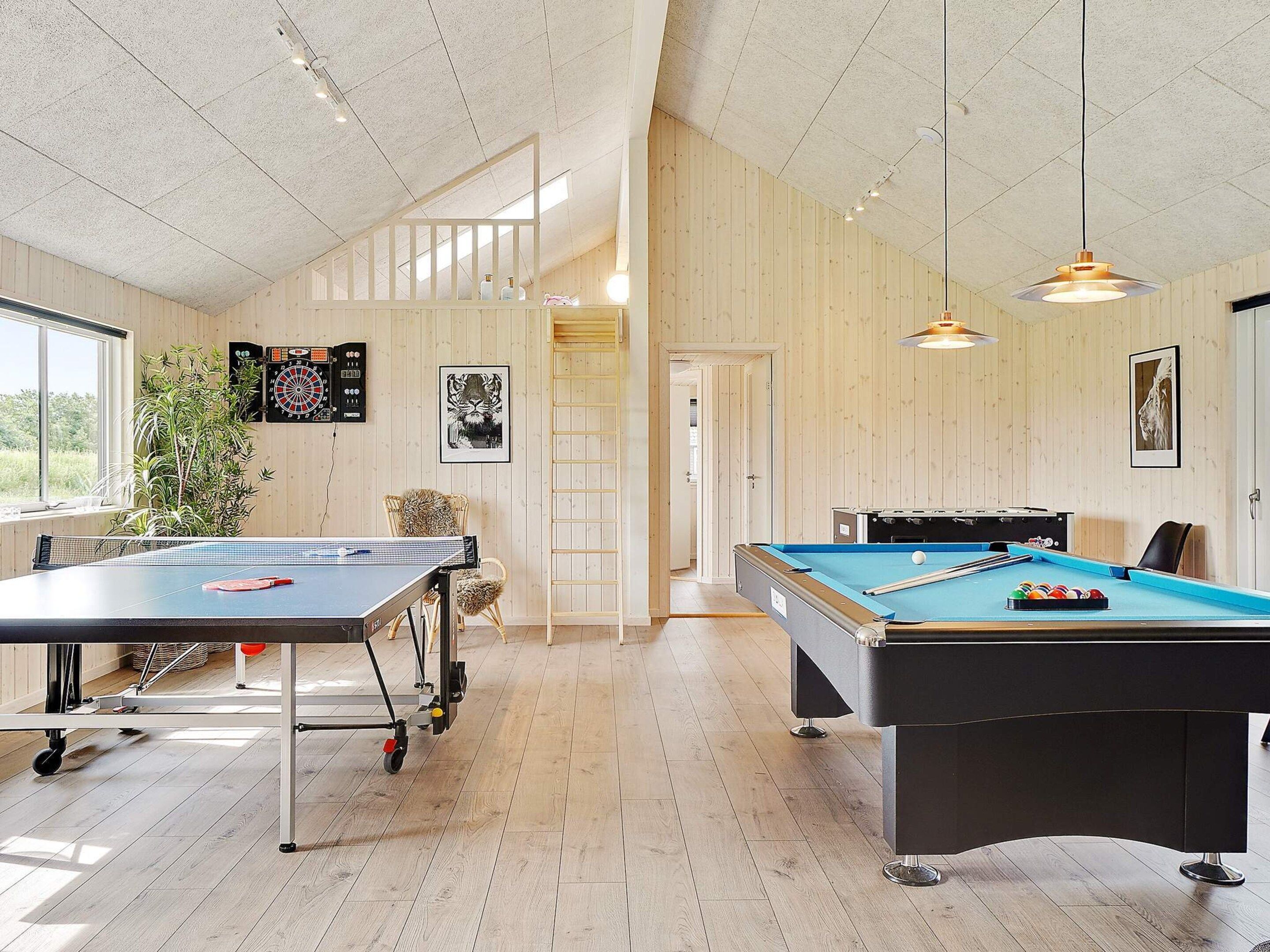 Une pièce équipée d’une table de ping-pong, d’un billard et d’un jeu de fléchettes. Une porte donne accès à une autre pièce, et on aperçoit une plante dans un coin.