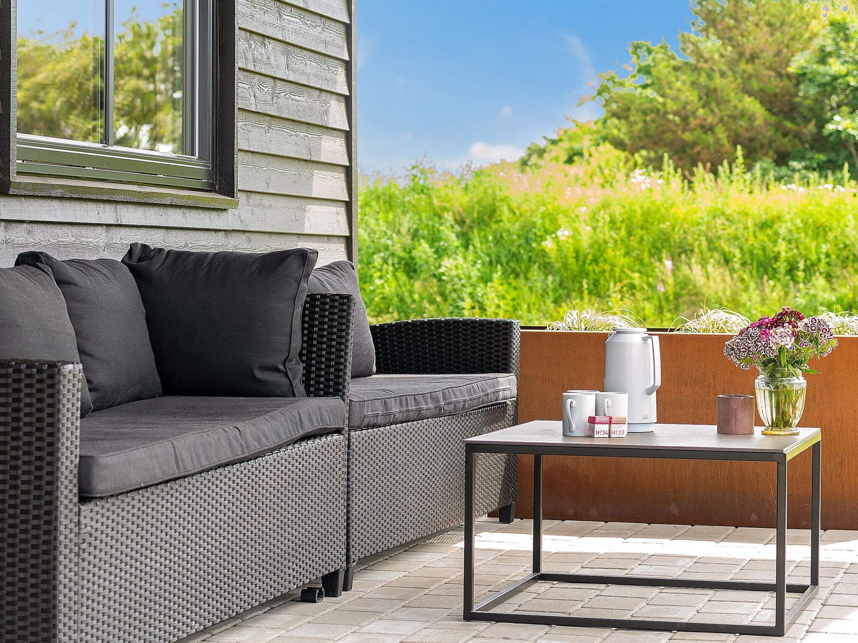 An outdoor seating area with a sofa and a table, a window, and a view of greenery.