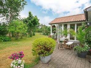 Relax in a serene garden setting with a patio area, featuring a table and chairs surrounded by lush greenery and flowering plants.