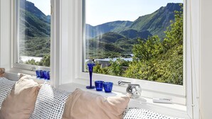 Une chambre dotée d’une grande fenêtre donnant sur des montagnes et un lac. On y trouve un lit avec des oreillers à motifs, une table de chevet sur laquelle reposent des verres bleus et une tasse en argent.