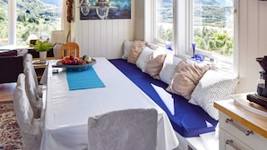 Une chambre avec un lit, une table à manger agrémentée d’un panier de fruits, une vue sur les montagnes par de grandes fenêtres et un tableau accroché au mur.