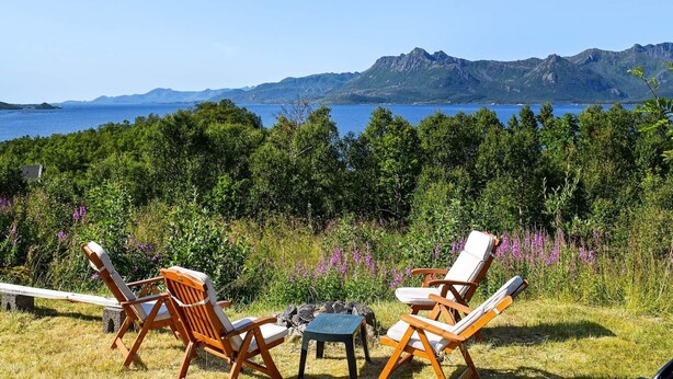 Un espace extérieur aménagé avec deux chaises en bois et une table, offrant une vue sur un plan d’eau et des montagnes.
