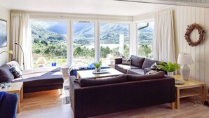 Un salon spacieux doté d’une grande fenêtre offrant une vue sur les montagnes, un coin salon confortable avec un canapé et des fauteuils, ainsi qu’une petite table avec une lampe.