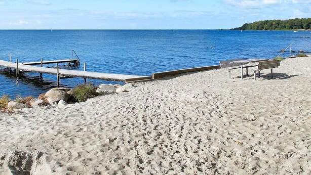 A beach with a wooden pier extending into calm blue water, a sandy shore, and a bench. - 4 Star Holiday Home in Nysted (Nysted)