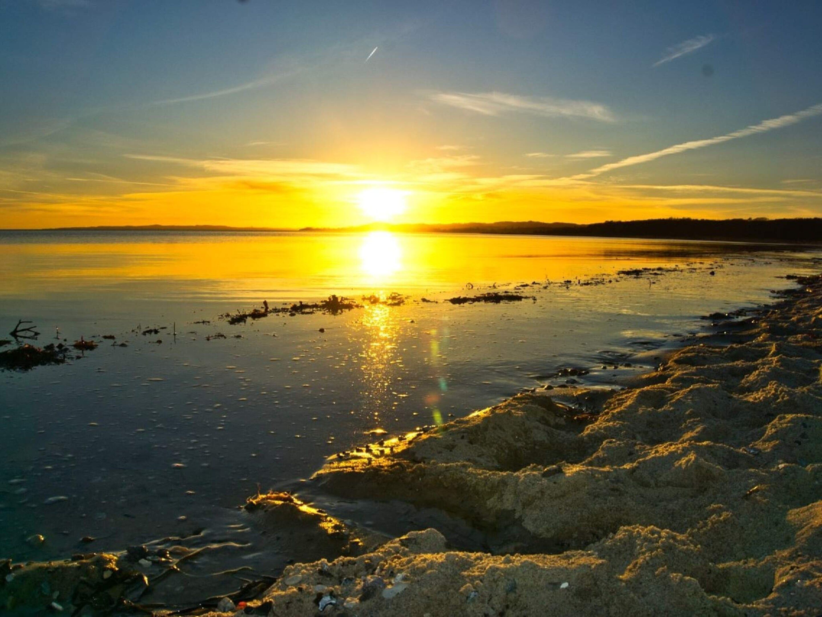 Un coucher de soleil sur une étendue d’eau calme, bordée d’une plage de sable.