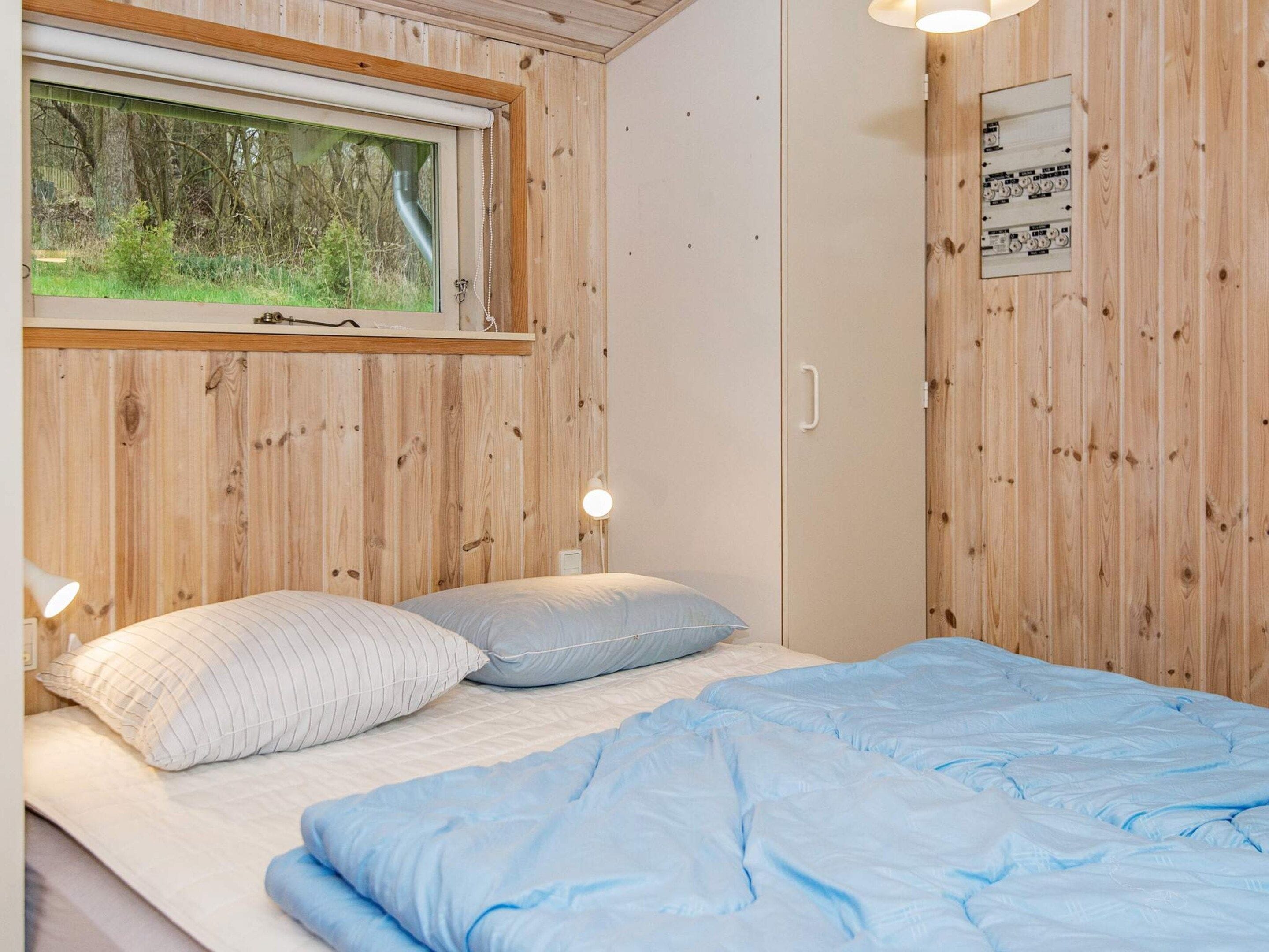 Een slaapkamer met houten paneelwanden, een bed met blauw beddengoed, een raam met uitzicht op groen, en een elektrisch paneel aan de muur.