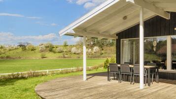 A covered outdoor area with a wooden deck, a table with chairs, and a view of a green field and trees.