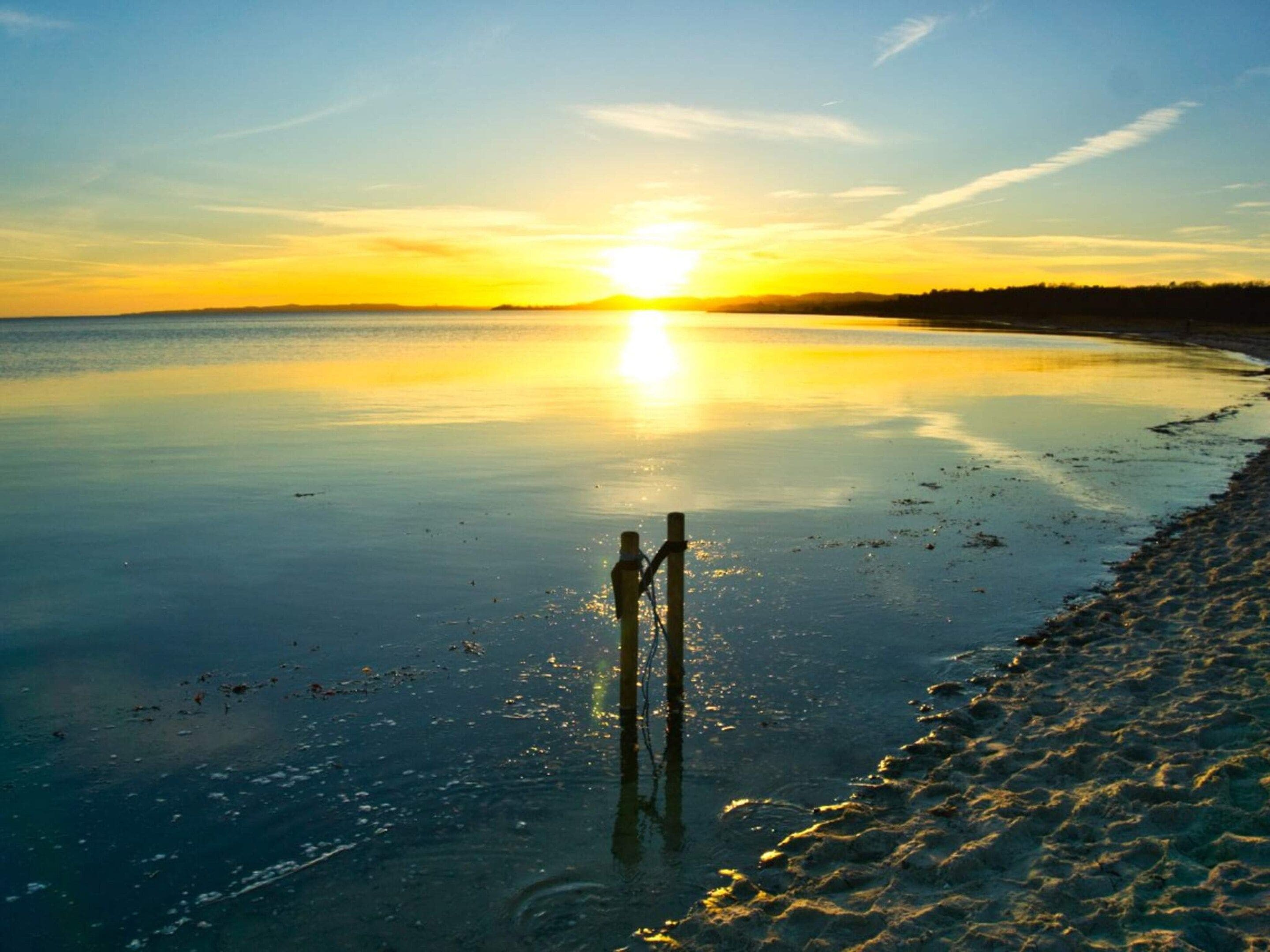 A calm body of water reflecting the sun, a wooden post, and a sandy beach.