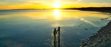 A calm body of water reflecting the sun, a wooden post, and a sandy beach.