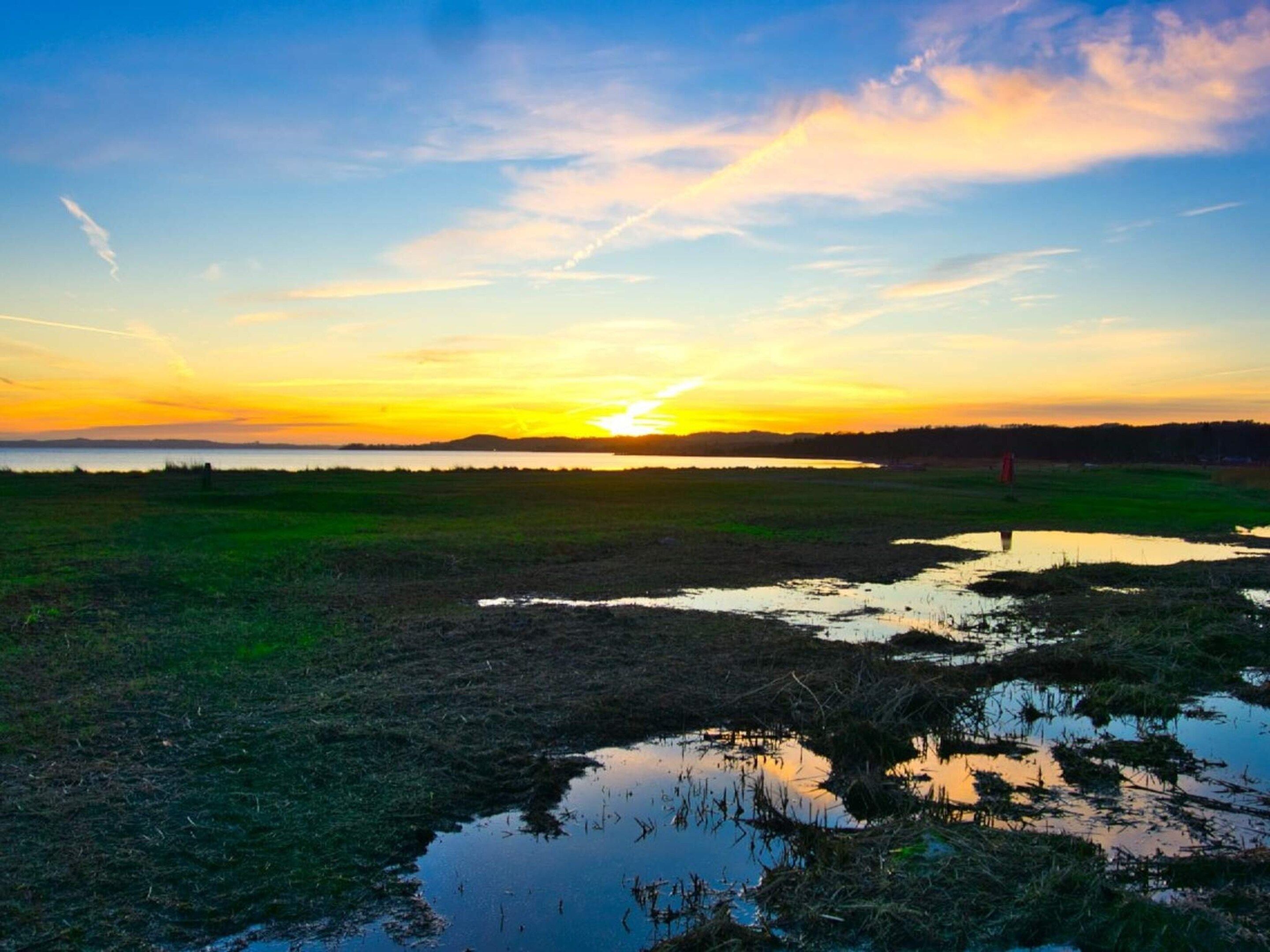 Eine flache Landschaft mit Sonnenuntergang, offenen Feldern und einem Gewässer, das den Himmel spiegelt.
