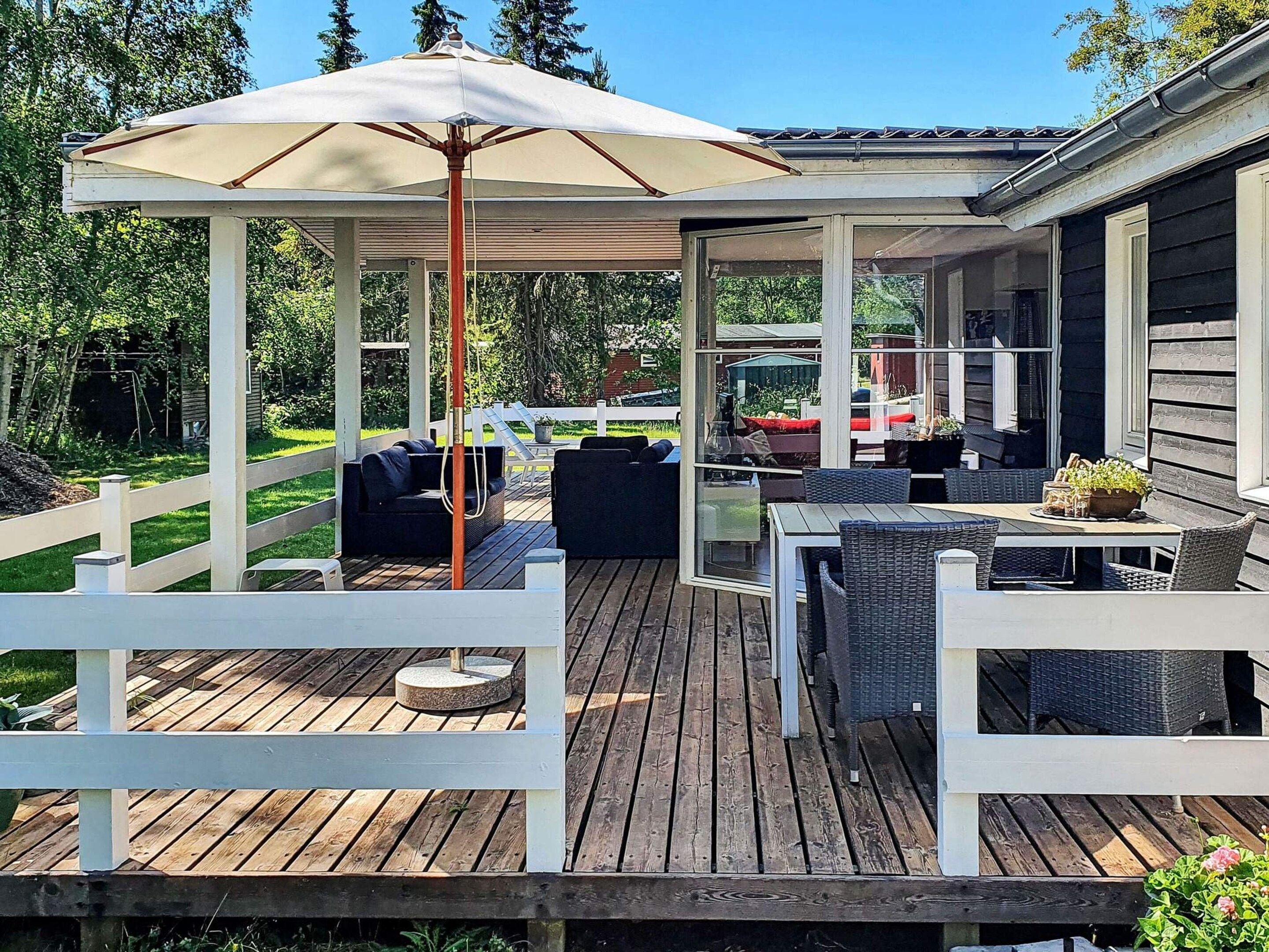 Een overdekt terras buiten met zitplaatsen en een tafel, omringd door een witte hekken en begroeiing.