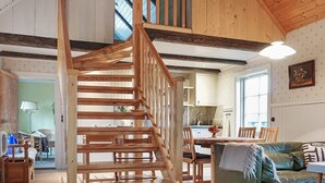 Eine Holztreppe führt zu einer oberen Ebene, einem Essbereich mit Tisch und Stühlen sowie einem Wohnzimmer mit Sofa und Lampe.