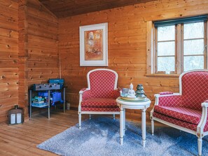 A cozy cabin interior with wooden walls, two red patterned chairs, a small table with decorative items, and a framed picture on the wall.