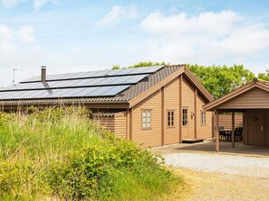 Een houten vakantiehuisje met een dak van zonnepanelen, een overdekt terras en een oprit met grind.