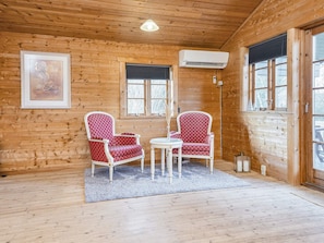 A wooden cabin interior with two red patterned chairs, a small round table, a framed picture on the wall, and an air conditioning unit.