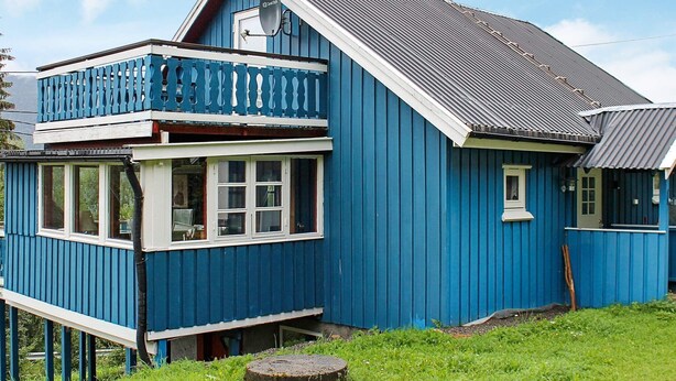A two-story blue wooden house with a balcony, a visible satellite dish, and a chimney.