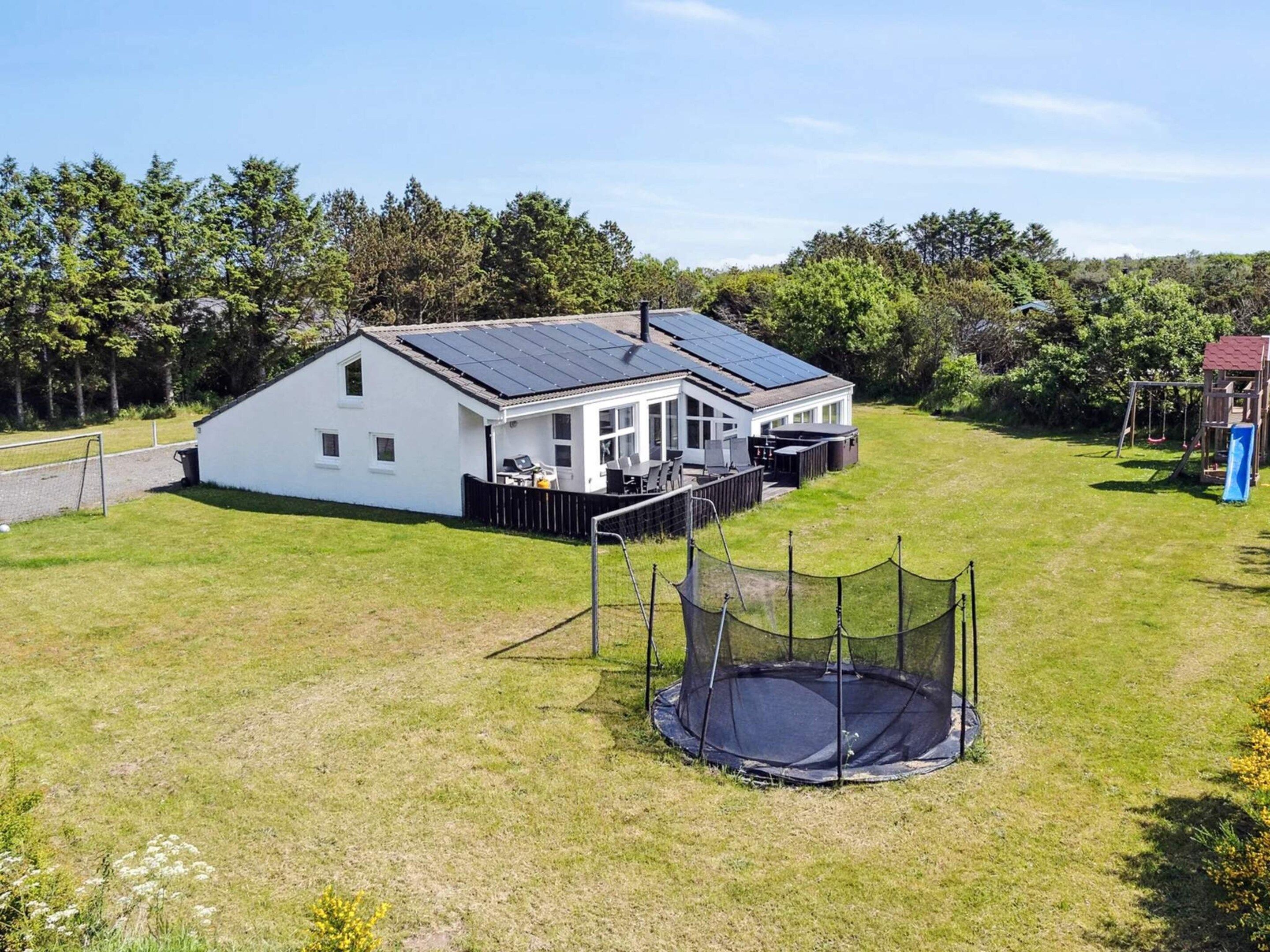 Una casa blanca con techo solar, un trampolín y un conjunto de columpios en un patio cubierto de césped.
