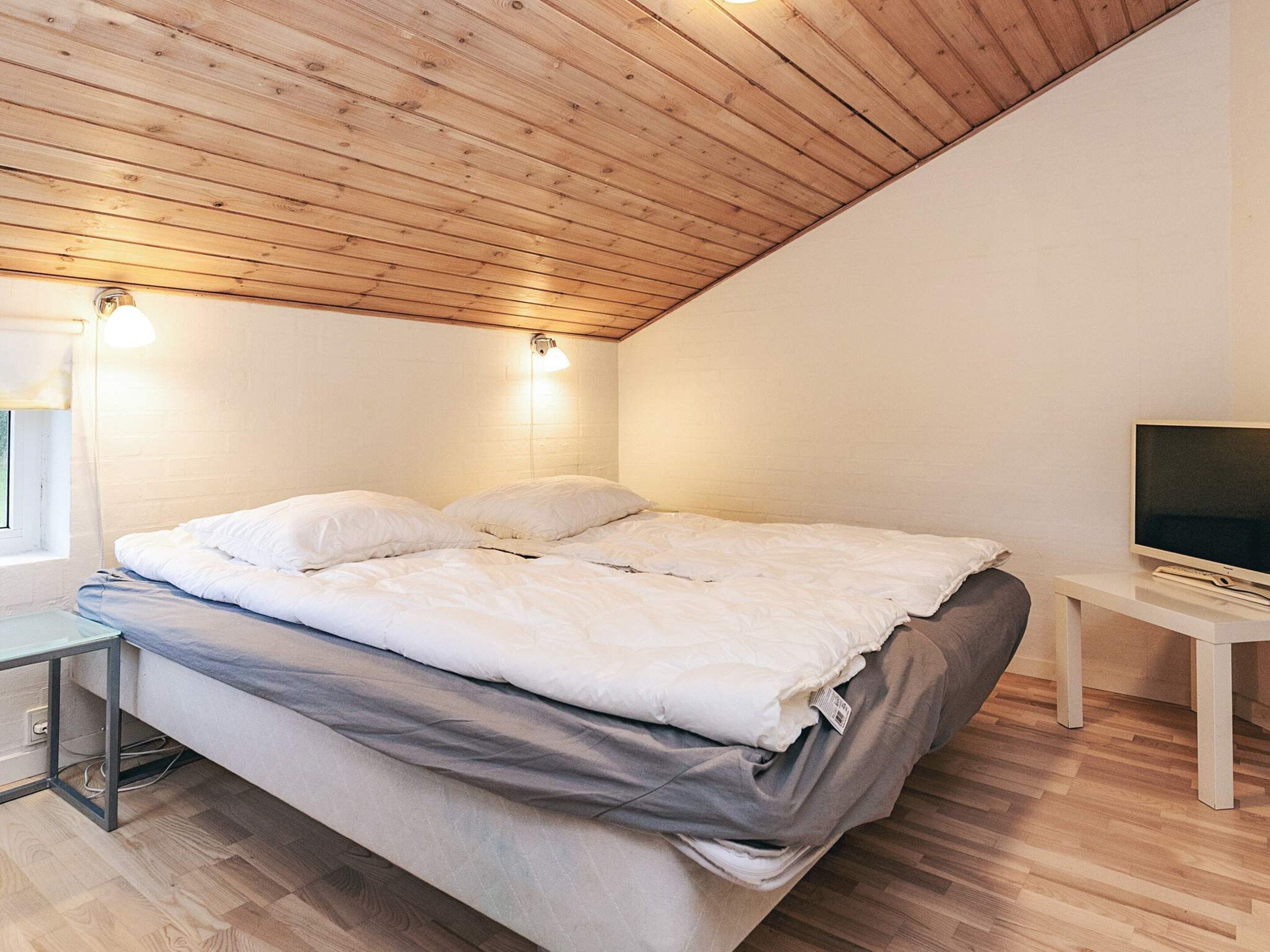 A bedroom with a wooden ceiling, a bed with white bedding, a small bedside table with a TV, and a window with a light fixture.