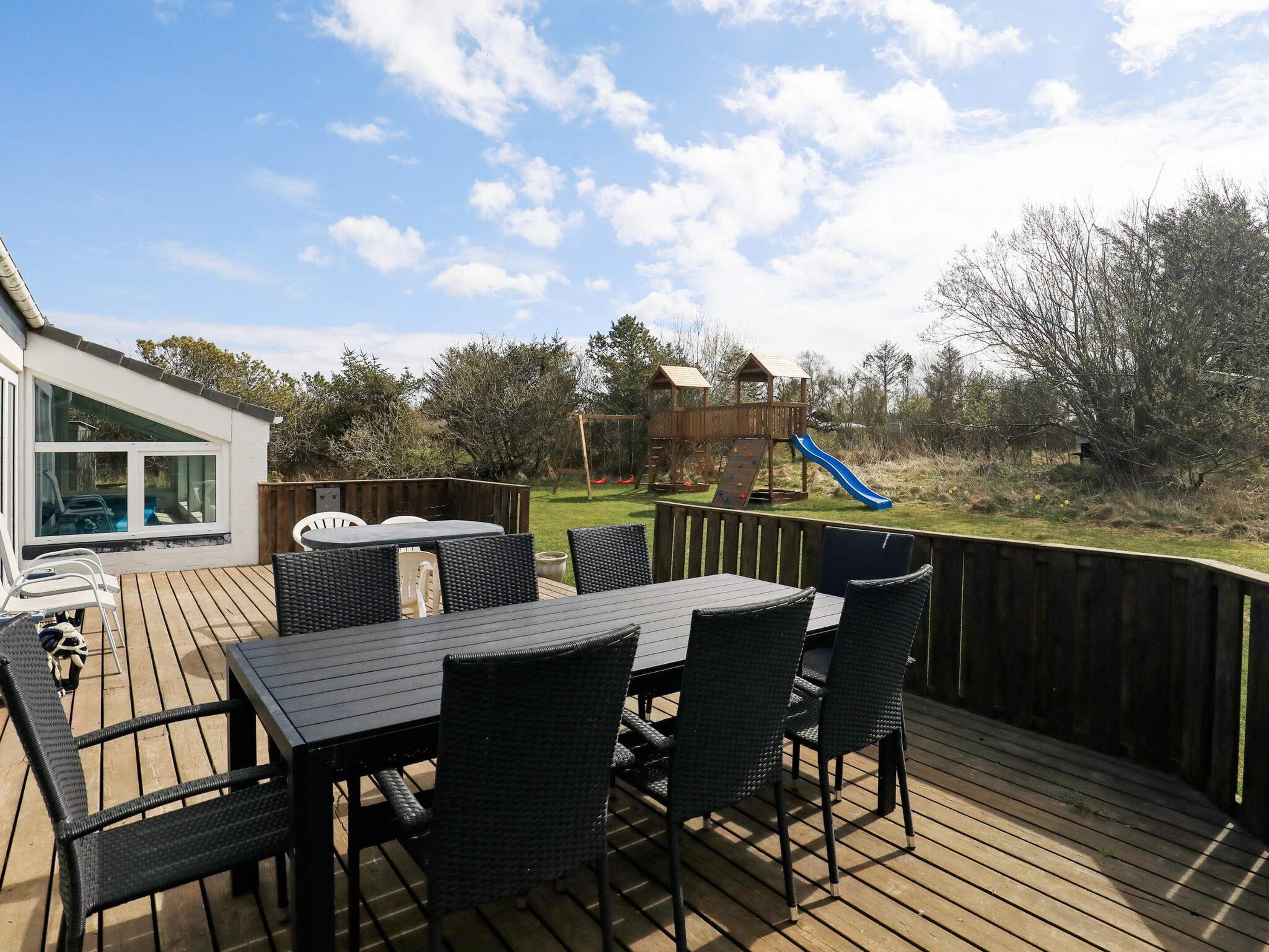 En udendørs terrasse med et bord og stole, et trælegeland med rutsjebane og udsigt over en græsplæne med træer.