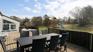 En udendørs terrasse med et bord og stole, et trælegeland med rutsjebane og udsigt over en græsplæne med træer.