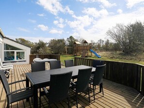 An outdoor deck with a table and chairs, a wooden play structure with a slide, and a view of a grassy area with trees. - 5 Star Holiday Home in Hjorring (Hjørring)