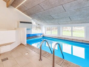 Een binnenzwembad met helderblauw water, een metalen zwemladder en uitzicht op het buitengebied door grote ramen.