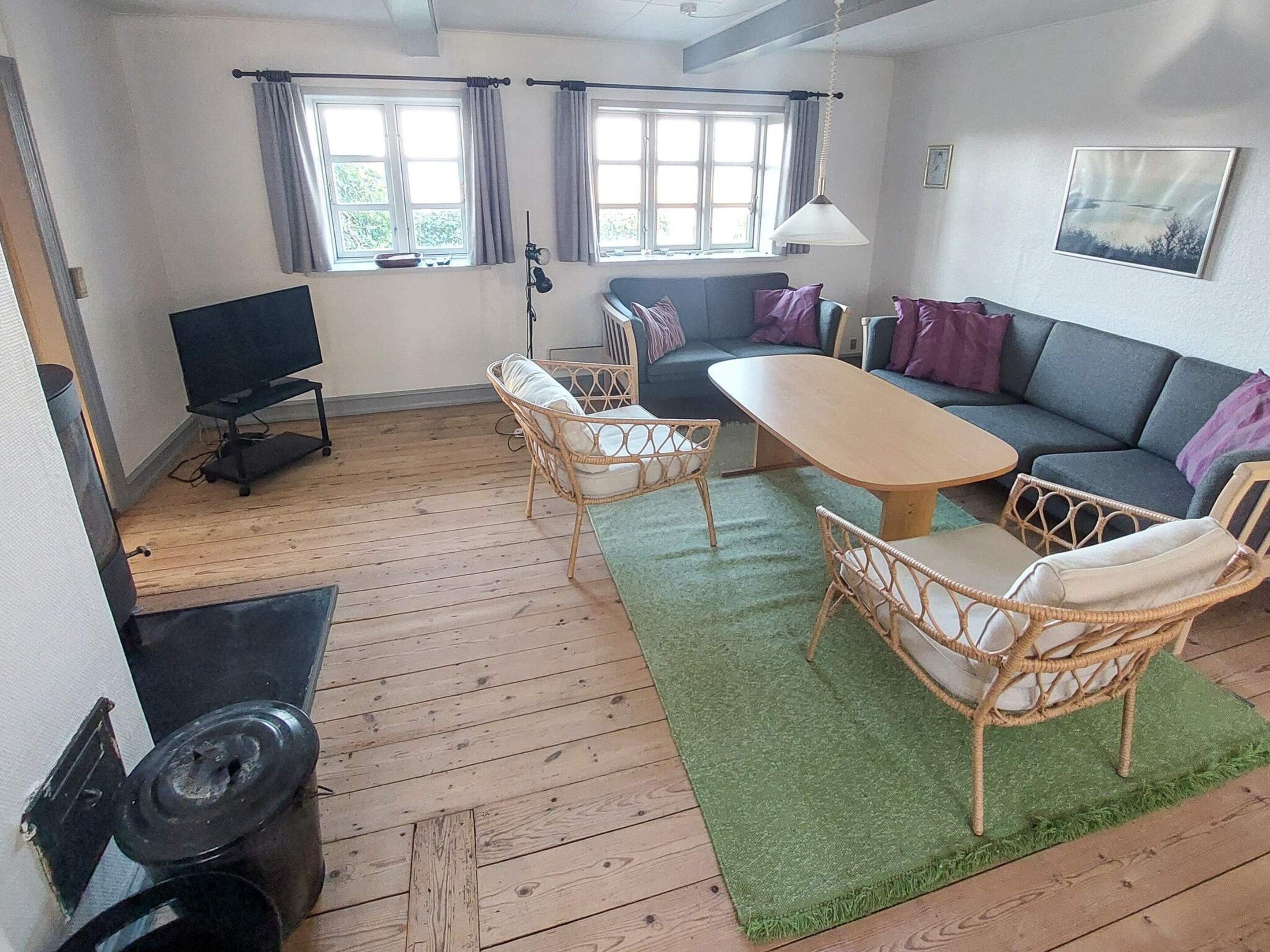 Et møbleret opholdsstue med sofa, to fletstole, et sofabord, et fjernsyn og et vindue med gardiner.