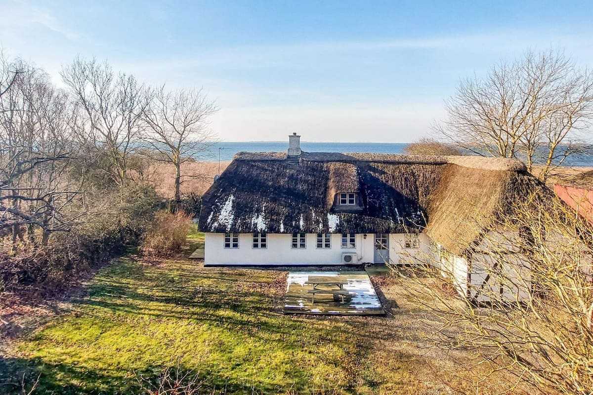 A white house with a thatched roof, surrounded by trees and a grassy area with a bench.