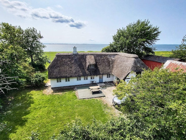 A white house with a thatched roof, surrounded by greenery and a view of the sea. - 8 Person Holiday Home in Svendborg (Svendborg)
