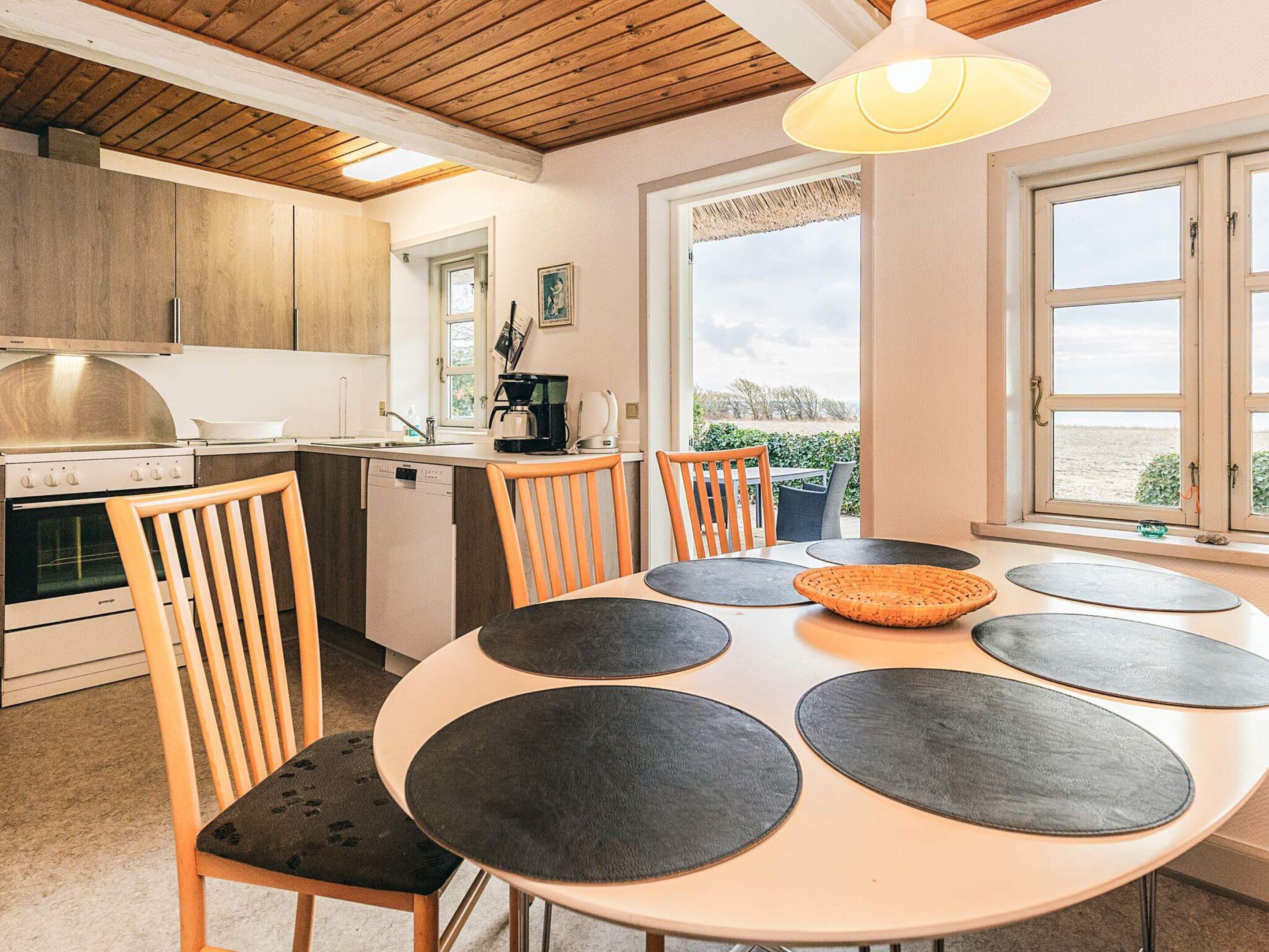 Et moderne køkken med træskabe, et spiseområde med et rundt bord og stole, samt udsigt gennem et stort vindue.