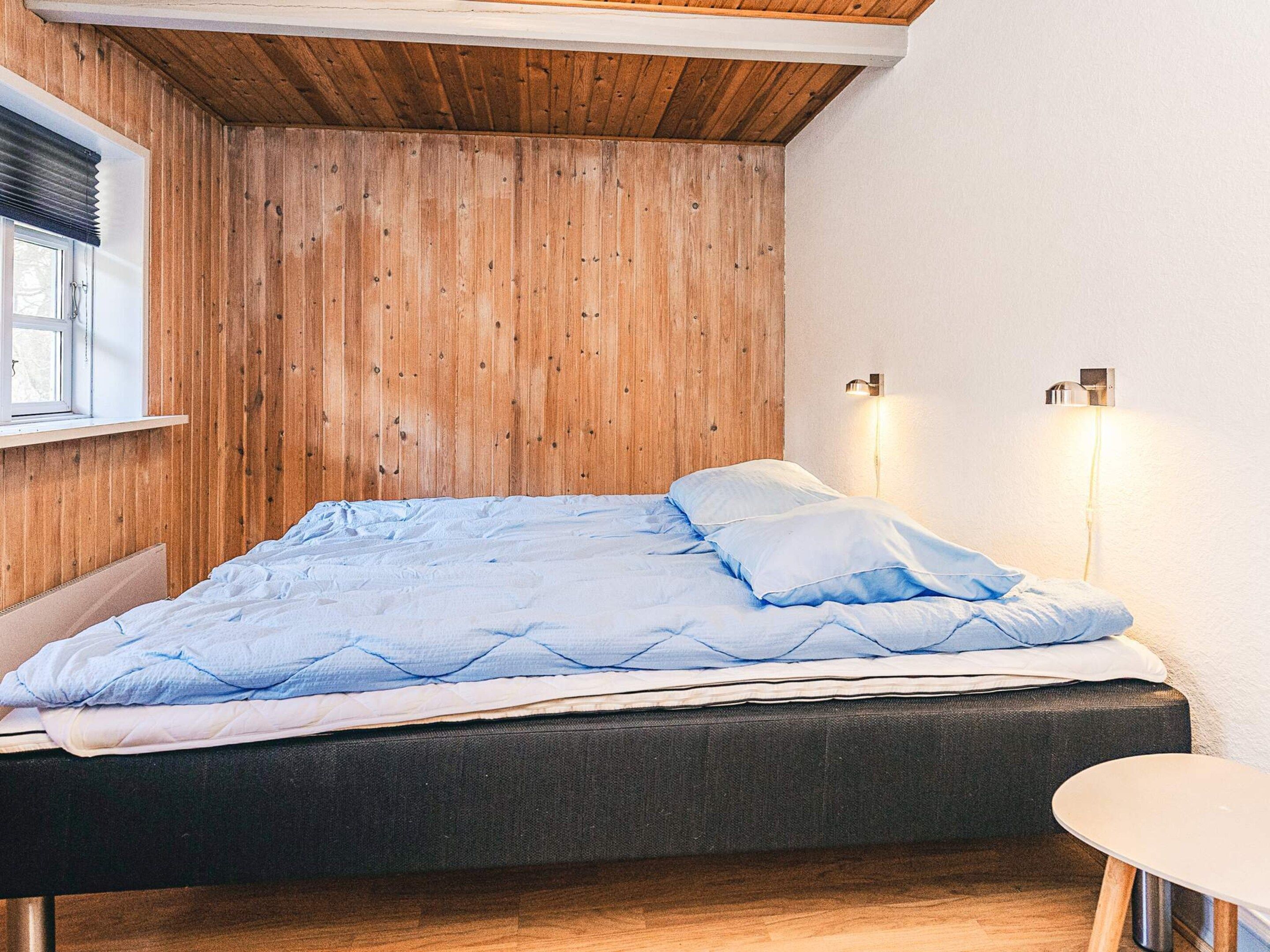 A bedroom with a wooden slatted ceiling, a bed with blue bedding, a bedside table, and two wall-mounted lights.