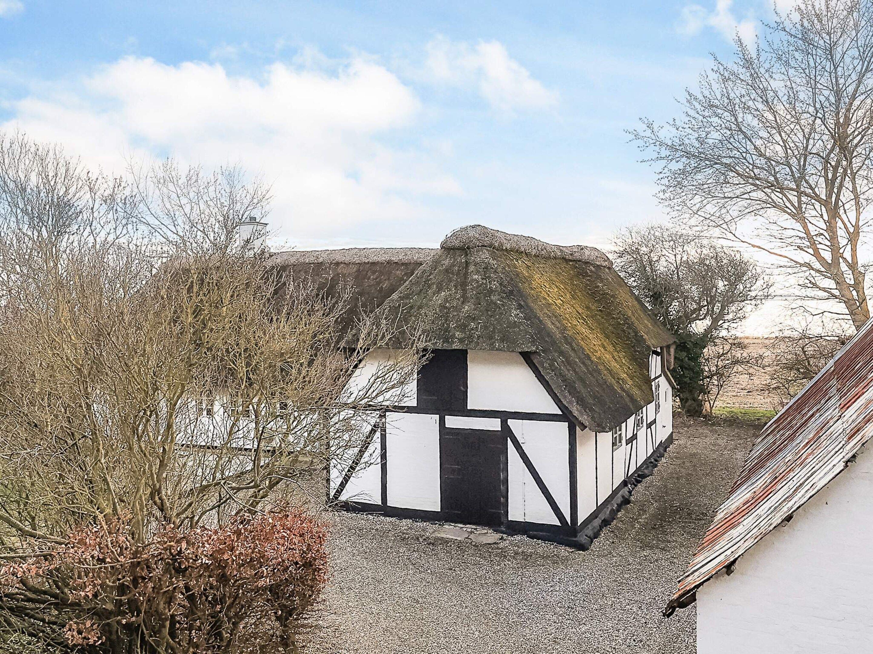 A traditional thatched-roof house with white and black timber framing, surrounded by trees and a gravel path.