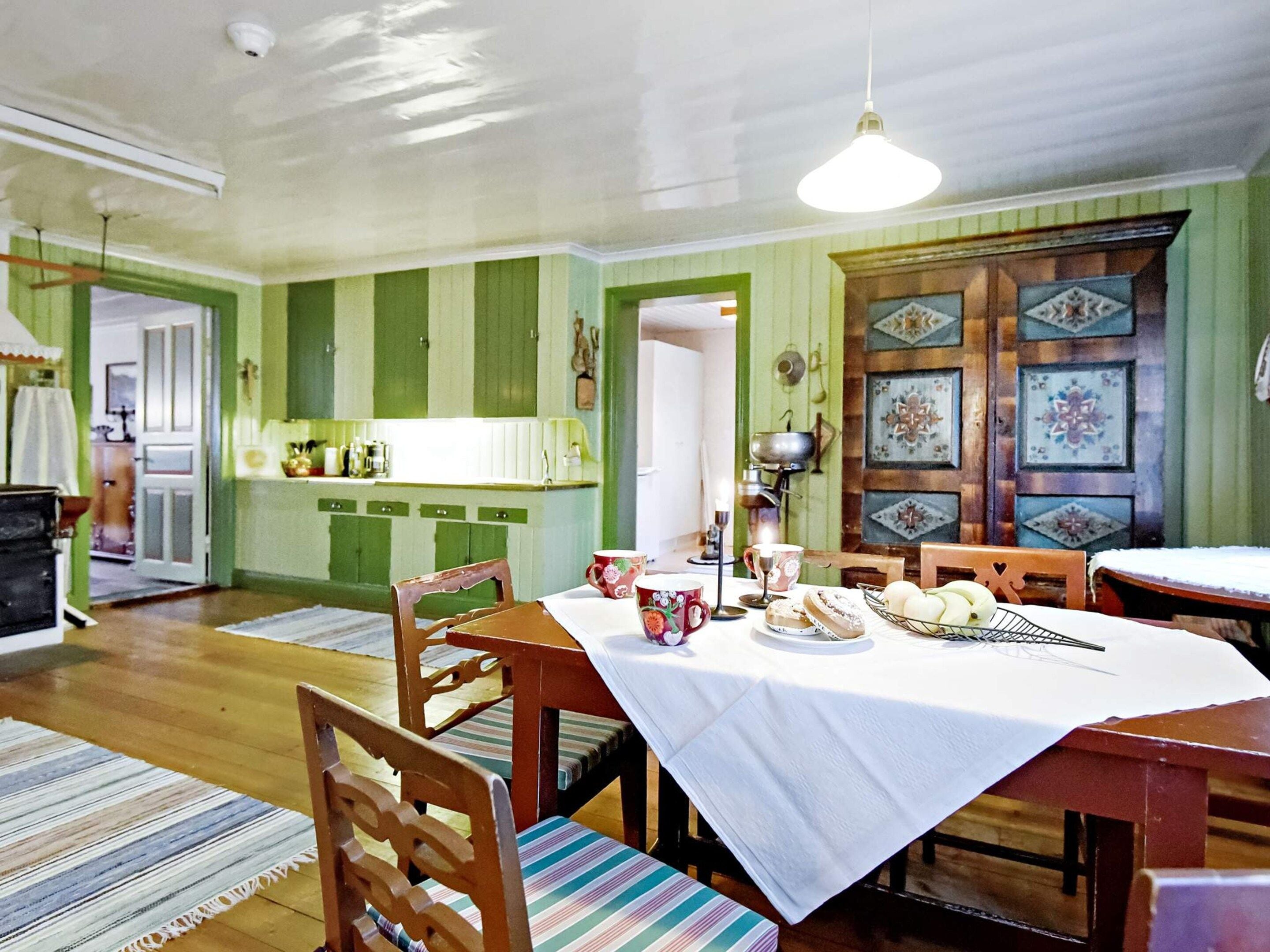 A dining area with a table set for four, chairs, a patterned rug, and a kitchen in the background.