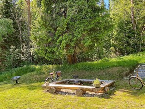 A rustic outdoor setting with a wooden fire pit surrounded by logs and stones, a bicycle, and a wheelbarrow.