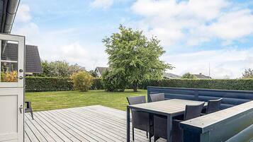 Una terraza con mesa y sillas, un árbol grande y un seto al fondo.