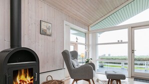 Een gezellige kamer met een houtkachel, een grijze fauteuil, een rieten mand en uitzicht op het landschap.
