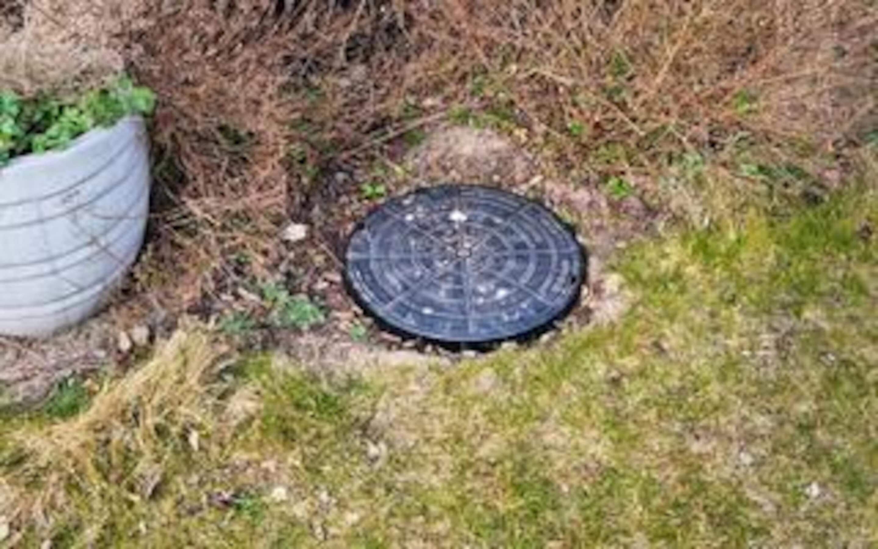 A manhole cover in a grassy area with a white planter nearby.