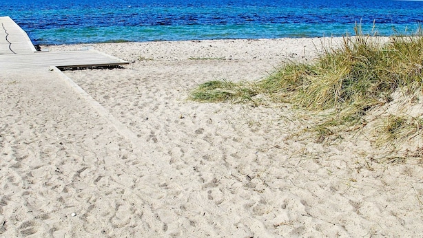 A sandy beach with a clear blue sea, a wooden deck, and some grass.