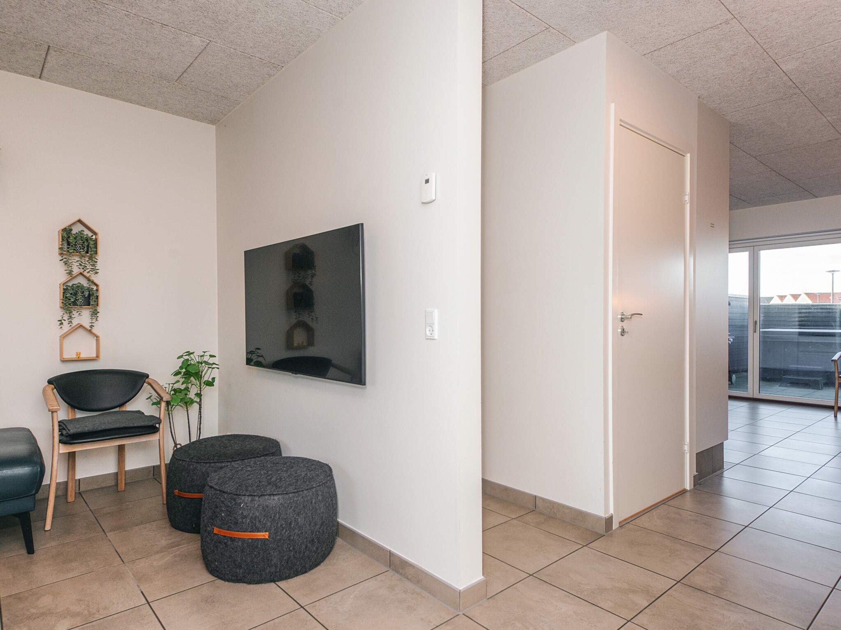 Un intérieur moderne avec un téléviseur à écran plat fixé au mur, un coin salon comprenant un fauteuil et un pouf, et une porte donnant sur un espace extérieur.