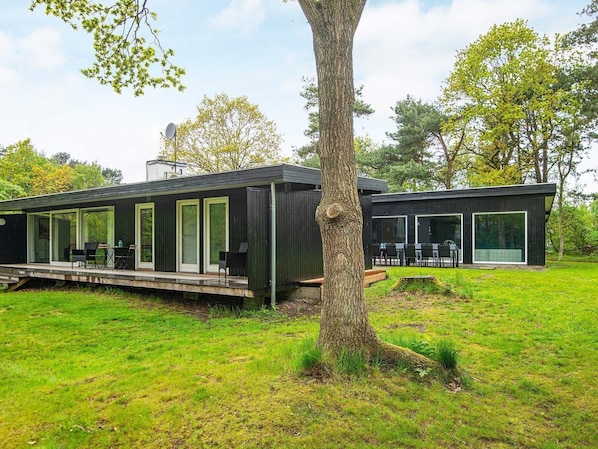 A modern house with a deck and a dining area, surrounded by a grassy yard and trees.