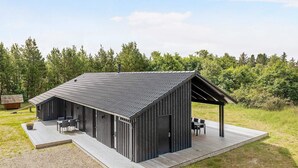 A modern, single-story building with a sloped roof, a covered outdoor seating area, and a gravel driveway. - Holiday Home in Jerup (Jerup)